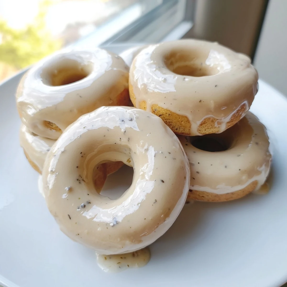 Freshly baked Earl Grey mochi donuts arranged on a white plate, their glossy tea-infused coating catching the light while a dusting of speckled tea leaves tops the sweet glaze.