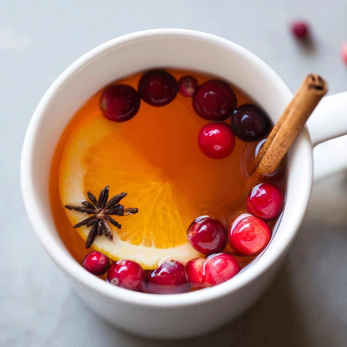 Steaming Orange Cranberry Tea spiced with cinnamon sticks and floating fresh cranberry slices in a clear glass teapot
