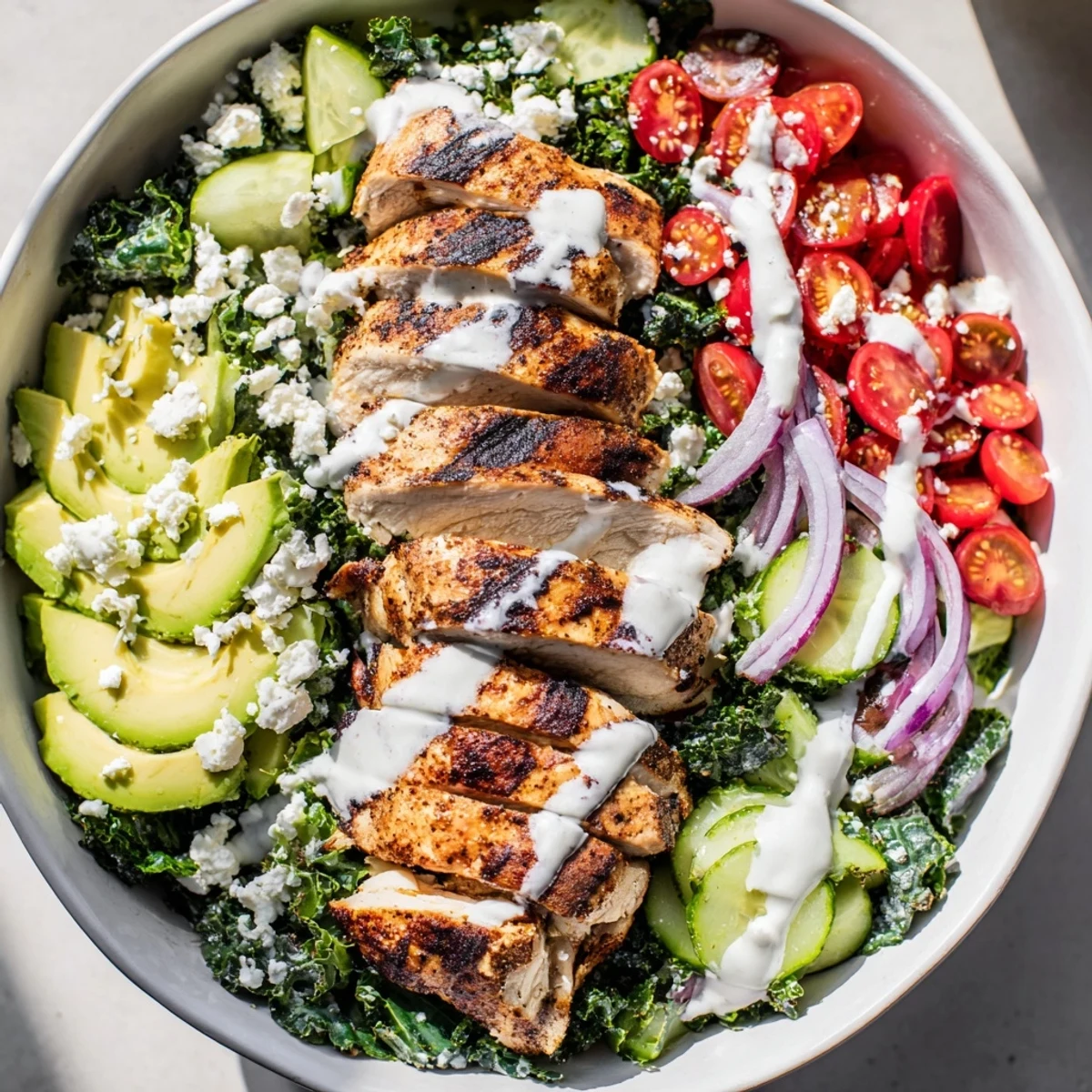 Sliced grilled chicken atop massaged kale with cherry tomatoes, cucumber, avocado, and crumbled feta